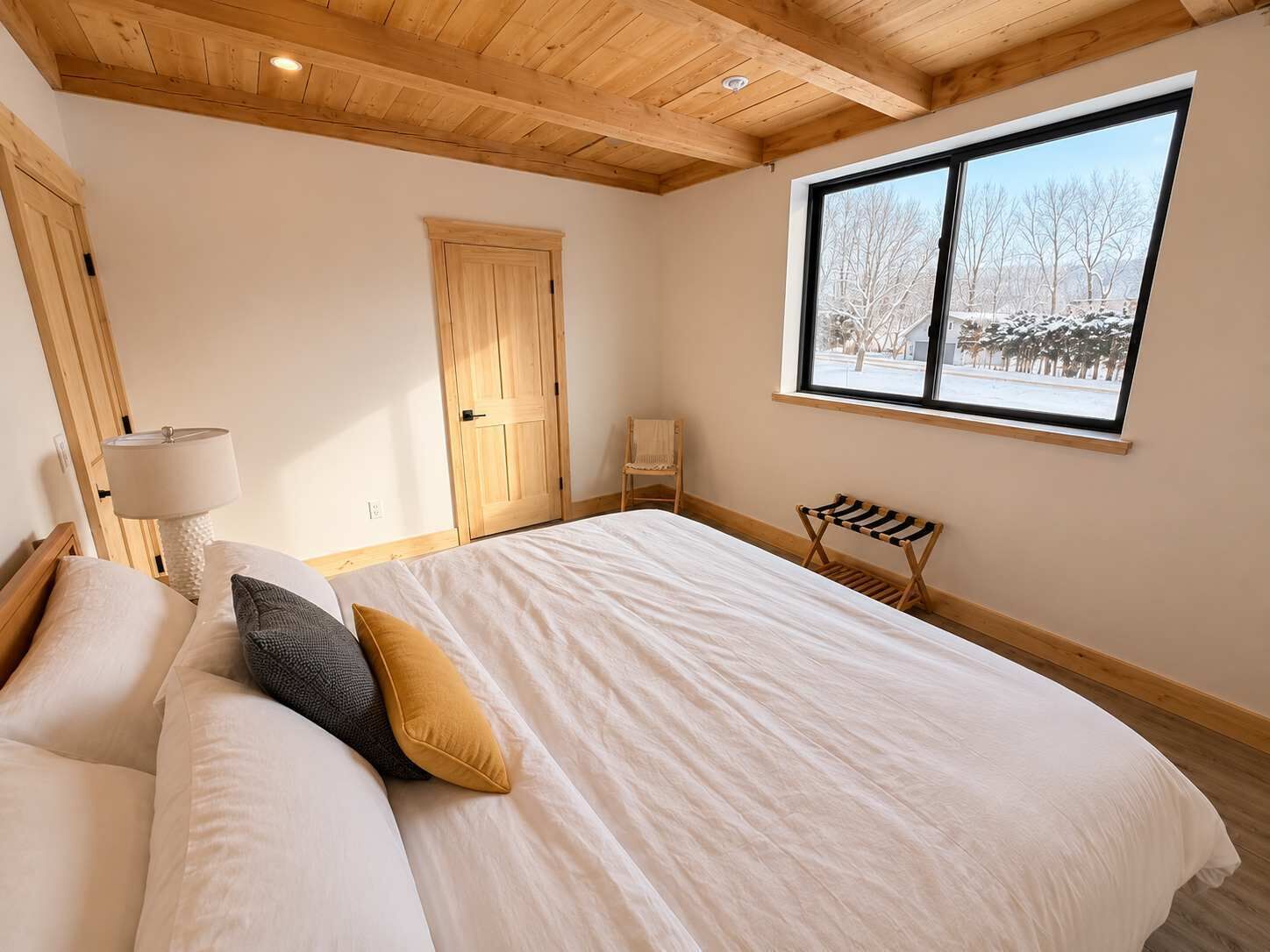 A bedroom with a large bed, a wooden ceiling, a window with a view of trees, and a door.