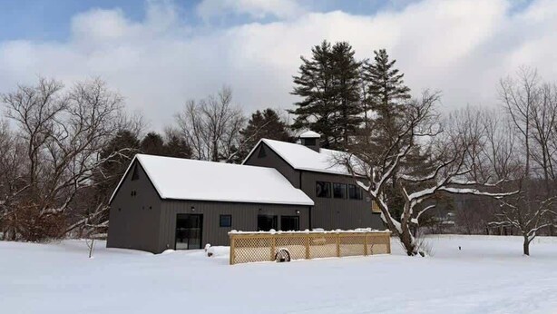 Un edificio de dos pisos con un techo cubierto de nieve, rodeado de árboles desnudos y un cielo despejado.