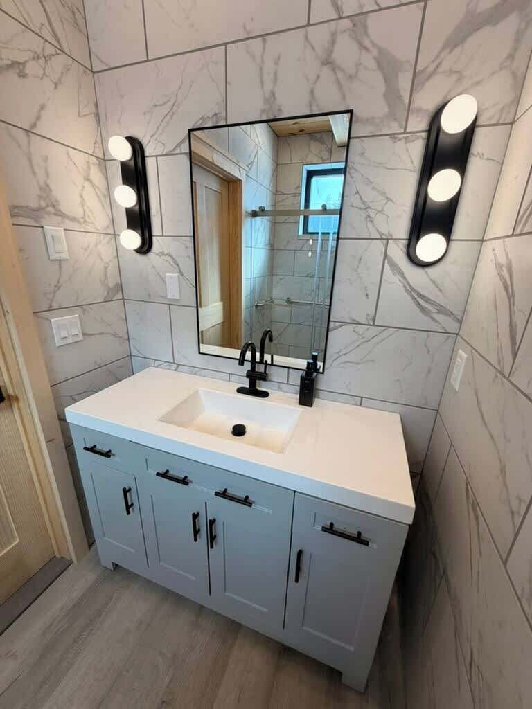 Un baño con pared de azulejos de mármol, un lavamanos blanco con grifo negro y un espejo con luces arriba.