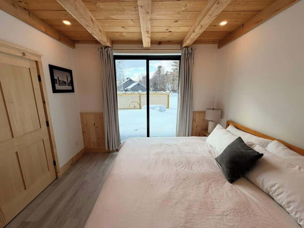 Un dormitorio con una cama grande, techo de madera, puerta corrediza de vidrio y vista a un paisaje nevado.