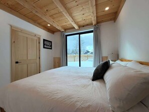 A bedroom with a large bed, wooden ceiling beams, a sliding glass door to a balcony, and a framed picture on the wall.