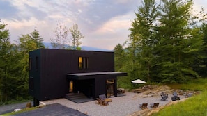 Una cabaña moderna negra con camino de grava, mobiliario exterior y rodeada de bosque.