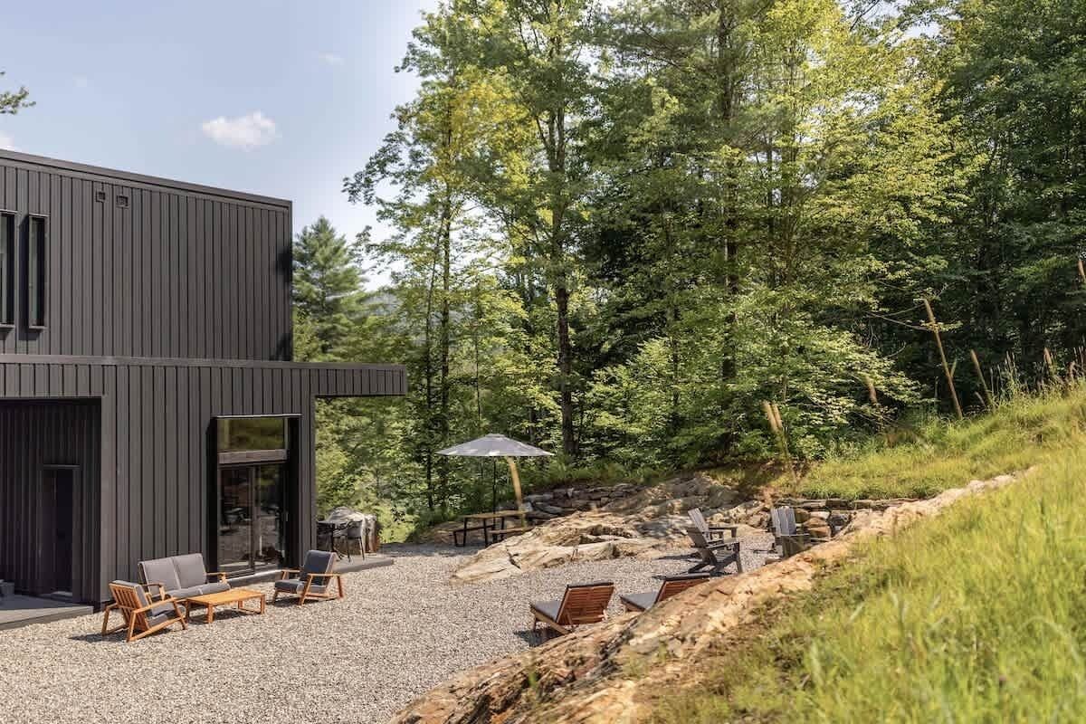 A modern building with a gravel patio, outdoor seating, and a forested backdrop.
