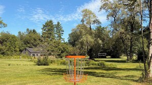 Ein grünes Feld mit einem Discgolf-Korb und einem Haus im Hintergrund.