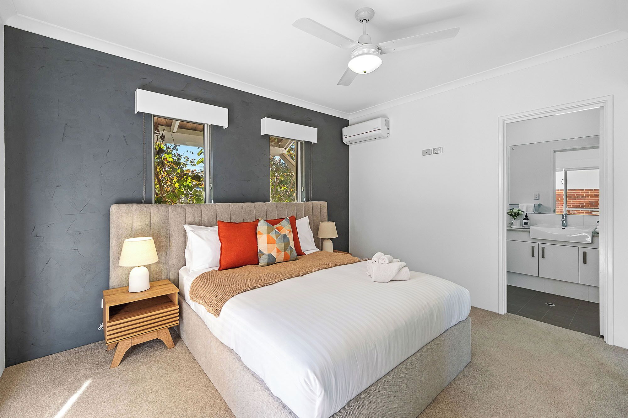 A modern bedroom with a large bed, a nightstand, a ceiling fan, and a view of the bathroom.