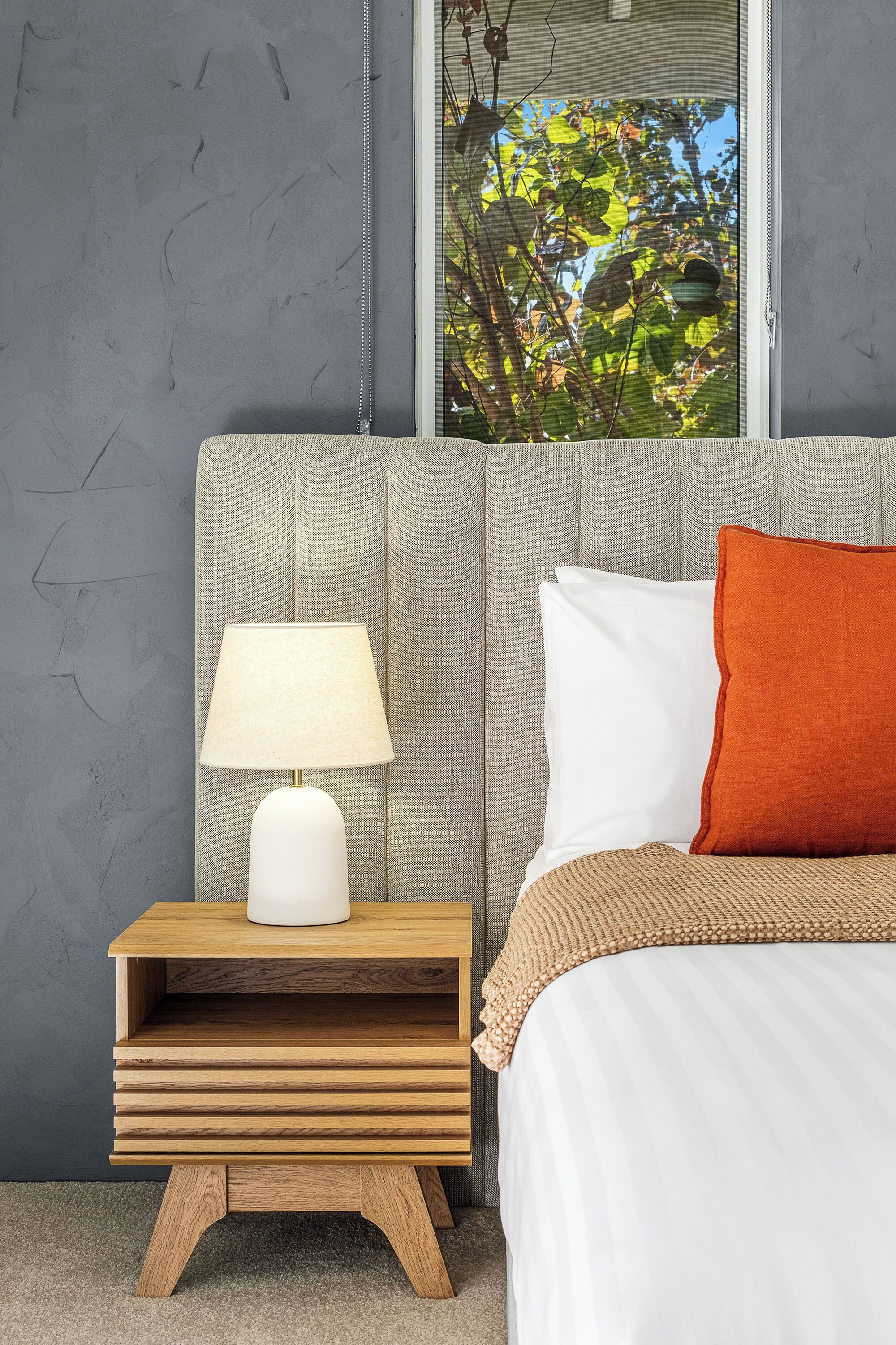 A bedroom with a bed, a nightstand with a lamp, a window with a view of trees, and a grey textured wall.