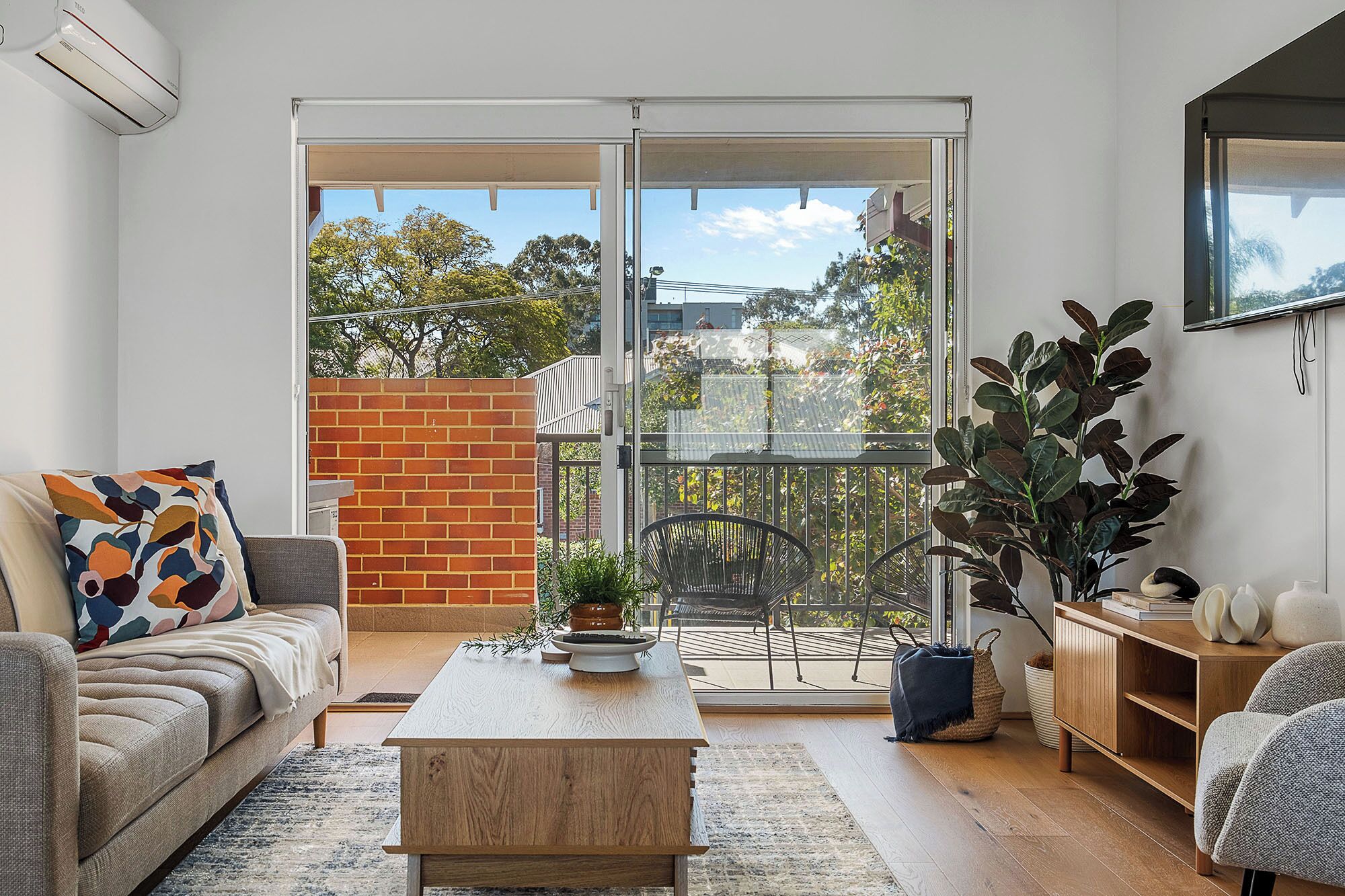 A modern living room with a sofa, a coffee table, and a balcony with a view of trees and buildings.