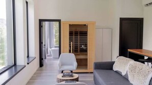 Ein modernes Wohnzimmer mit einem grauen Sofa, einem weißen Sessel und einem kleinen runden Tisch mit einem Buch. Ein Holzschrank mit Glastür ist zu sehen, im Hintergrund eine dunkle Tür.