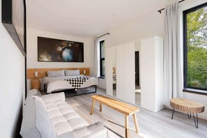 Un dormitorio moderno con una cama grande, un sofá, un banco de madera y vistas a la vegetación a través de una ventana.