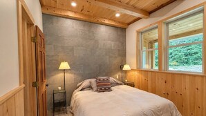 Un dormitorio con cama, dos lámparas de noche, un ventanal con vistas a follaje y paneles de madera.
