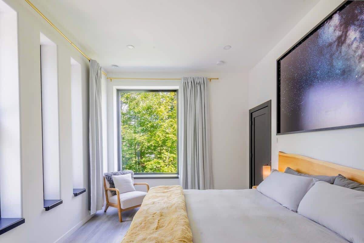 A modern bedroom with a large bed, a chair, a window with curtains, and a wall-mounted TV.