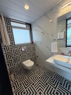 A bathroom with a geometric patterned floor, a white toilet, a sink with a mirror, and a window with a towel hanging on the wall.
