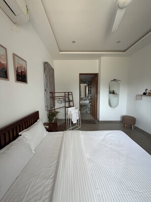 A hotel room with a large bed, a wooden headboard, a wall-mounted air conditioner, a mirror, and a small stool.