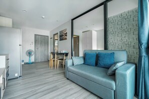 A modern living room with a grey sofa, a dining area with a table and chairs, and a large window with a patterned curtain. - The Title Residencies Naiyang Beach Apartment G507 (Thalang/Sakhu)