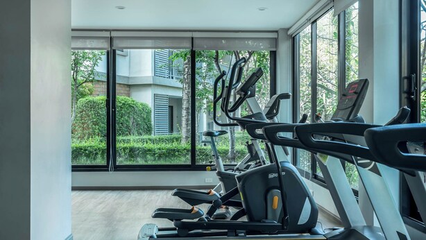Un gimnasio con equipamiento de ejercicio y grandes ventanales que ofrecen vistas a zonas verdes y edificios.