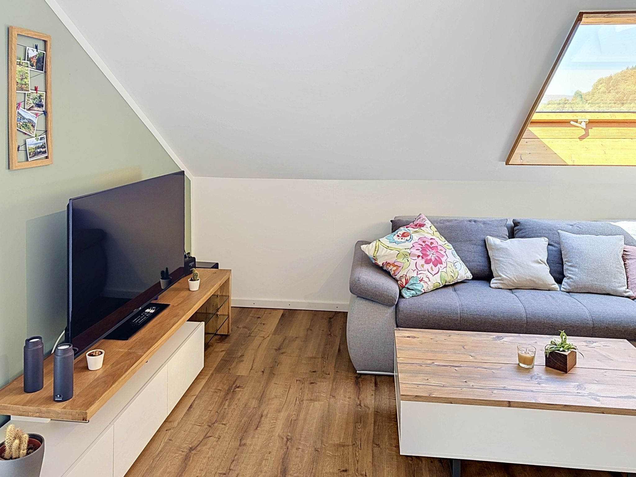Ein modernes Wohnzimmer mit einem Flachbildfernseher, einem grauen Sofa mit floralen Kissen, einem Holztisch und einer kleinen Topfpflanze.