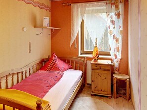 A small bedroom with a single bed, a wooden nightstand, a window with a curtain, and a wall-mounted shelf.