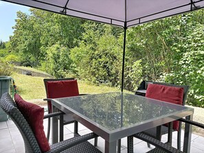 An outdoor patio with a glass-top table and wicker chairs, surrounded by a covered area and lush greenery.