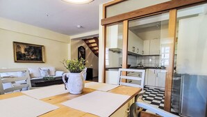 Un comedor con una mesa de madera, individuales blancos y una planta en maceta. Se ve una cocina al fondo, visible a través de una puerta de cristal.