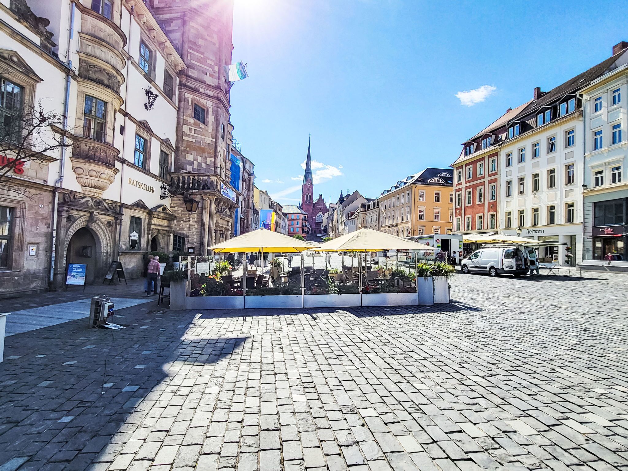 Ett torg med kullerstenar, omgivet av historiska byggnader, ett kyrktorn och uteserveringar med parasoller.
