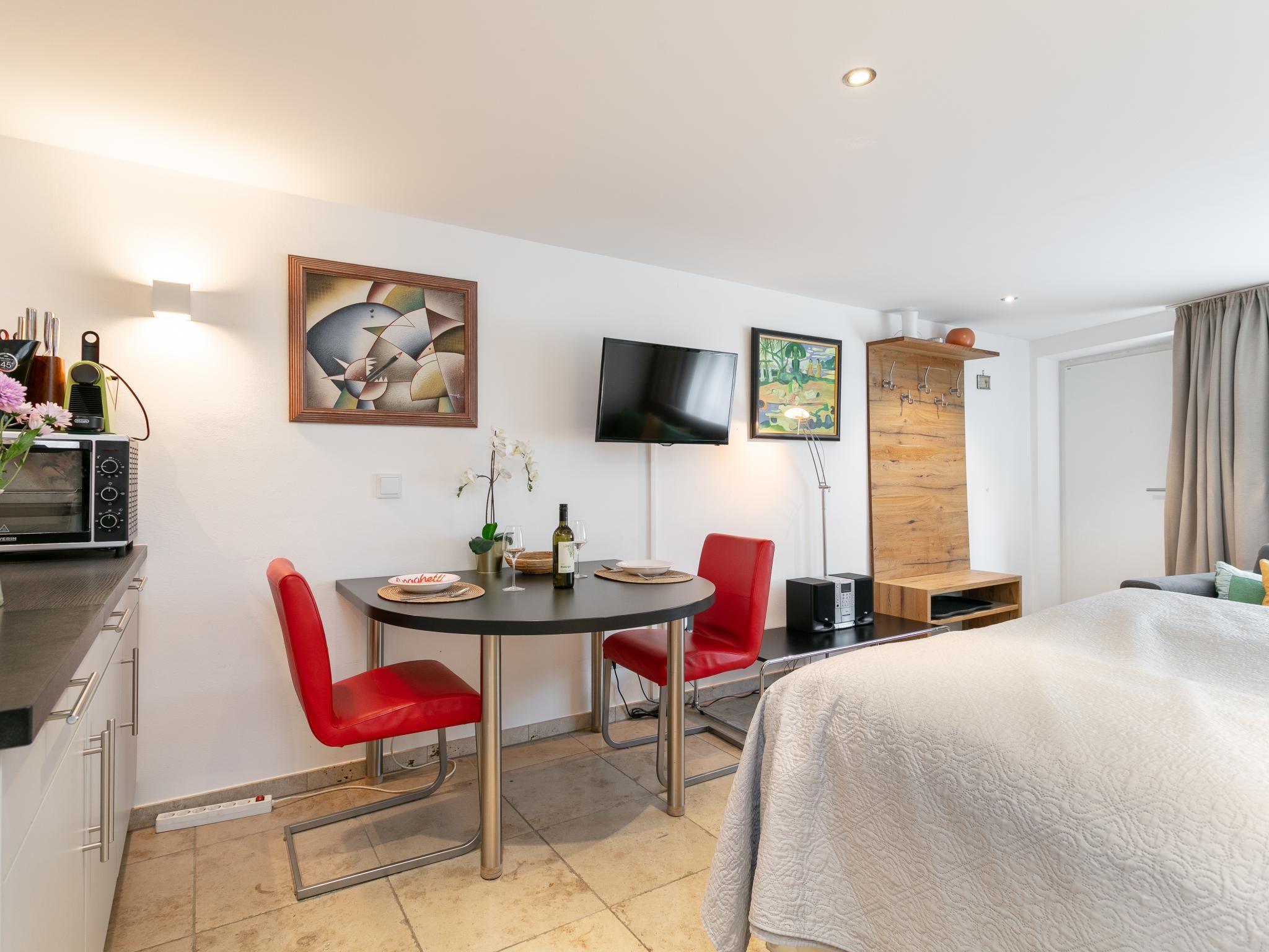 A modern hotel room with a bed, a dining table with chairs, a TV mounted on the wall, and artwork.