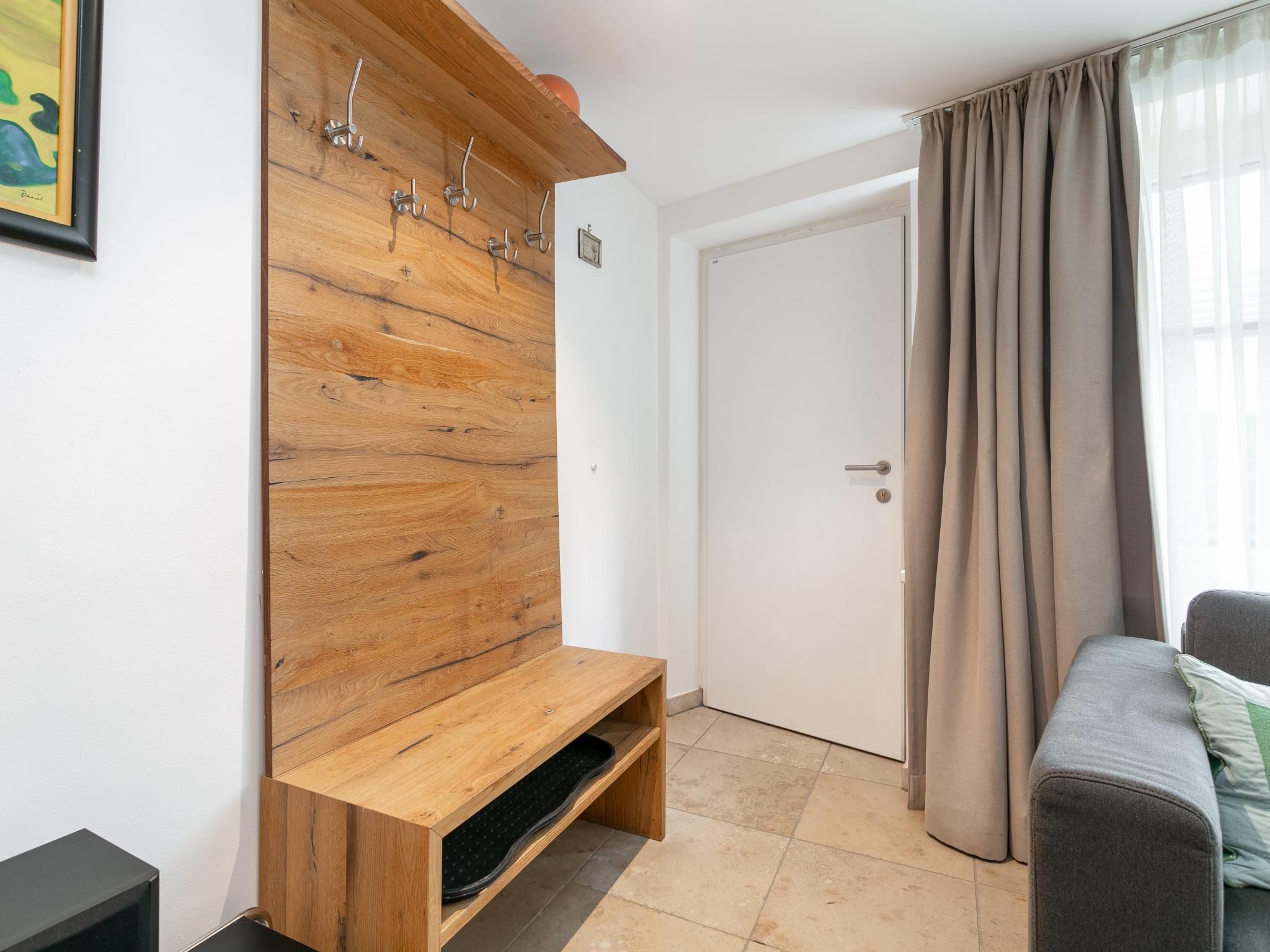 Een kamer met een houten kapstok aan de muur, een deur, een grijze bank en een raam met gordijnen.