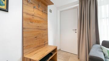 Een kamer met een houten kapstok aan de muur, een deur, een grijze bank en een raam met gordijnen.