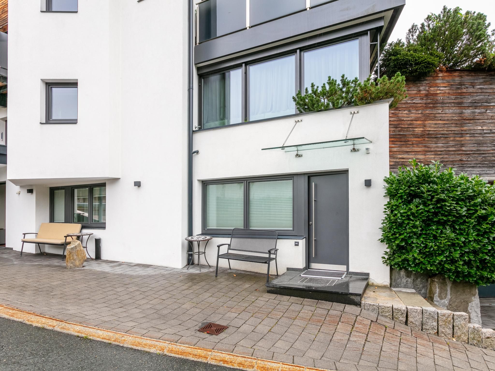 Een modern woongebouw met twee verdiepingen, een witte gevel, grote ramen en een klein terras met een bankje en een tafel.