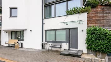 Een modern woongebouw met twee verdiepingen, een witte gevel, grote ramen en een klein terras met een bankje en een tafel.