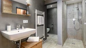 Ein modernes Badezimmer mit einem weißen Waschbecken, einem Holz-Waschtisch, einem Handtuchhalter, einem Spiegel, einer Duschkabine und einer Toilette.