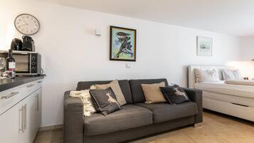 Un espace de vie moderne comprenant un canapé gris, un lit blanc, un coin cuisine avec un four à micro-ondes et une horloge murale.