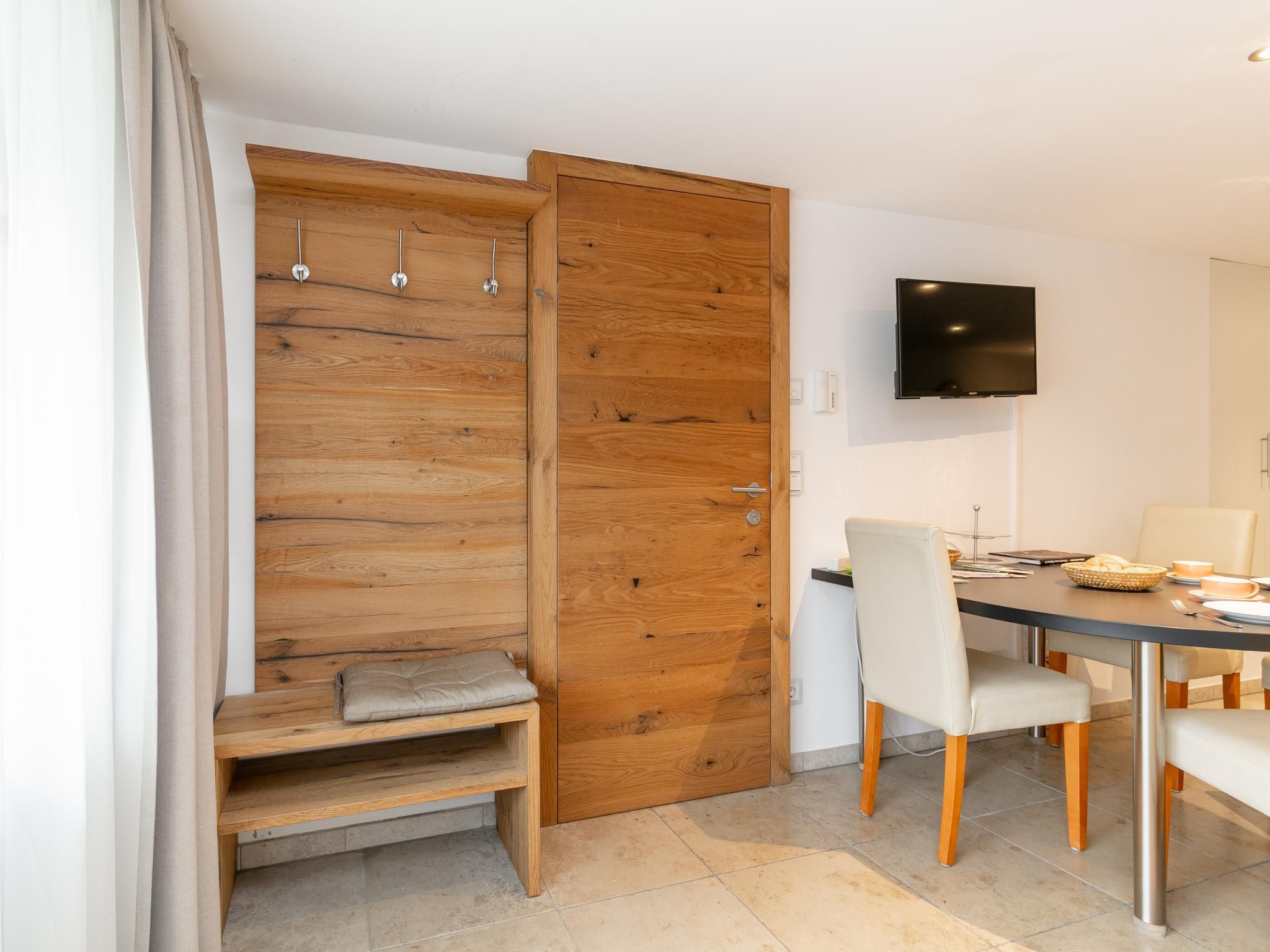 Une chambre moderne avec une armoire en bois, un banc, une table à manger avec des chaises et un téléviseur à écran plat fixé au mur.