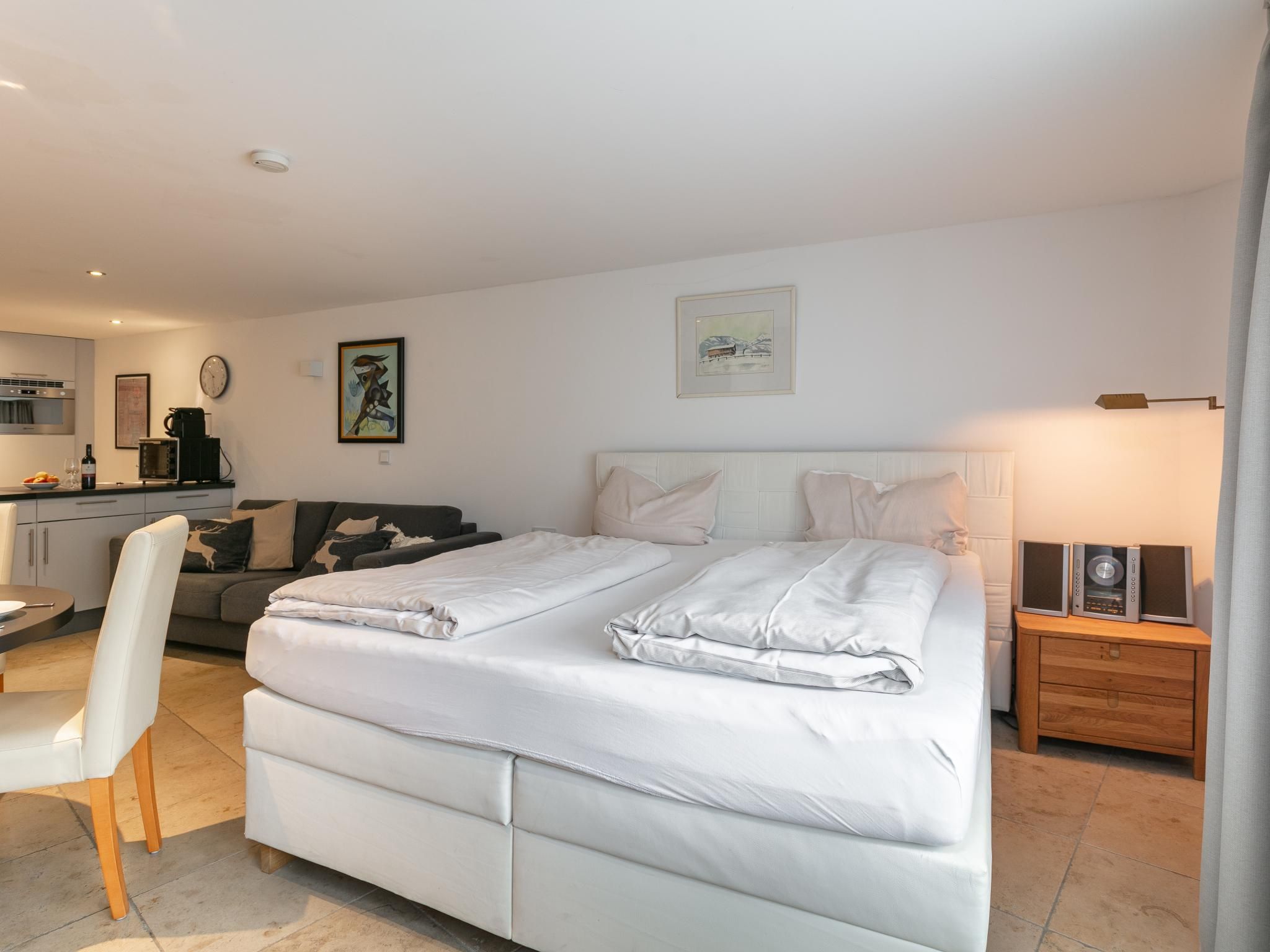 A modern bedroom with a large bed, a bedside table with speakers, a sofa, and a dining area with a table and chairs.