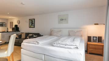 A modern bedroom with a large bed, a bedside table with speakers, a sofa, and a dining area with a table and chairs.