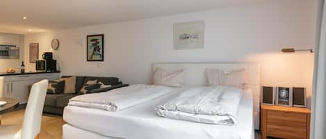 A modern bedroom with a large bed, a bedside table with speakers, a sofa, and a dining area with a table and chairs.
