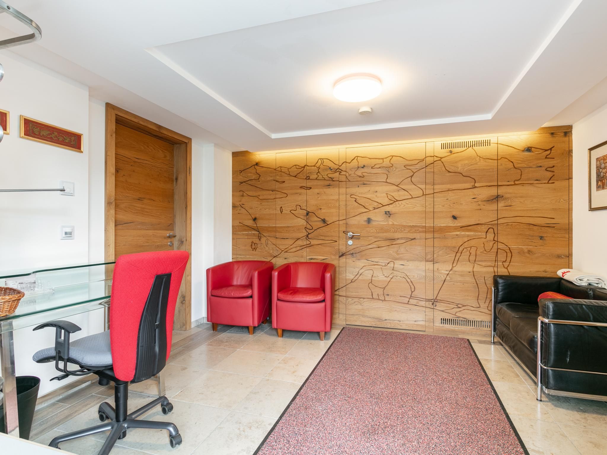Een modern receptiegebied met een houten wandpaneel, een glazen toonbank, rode stoelen, een zwarte leren bank en een rode vloerkleed.
