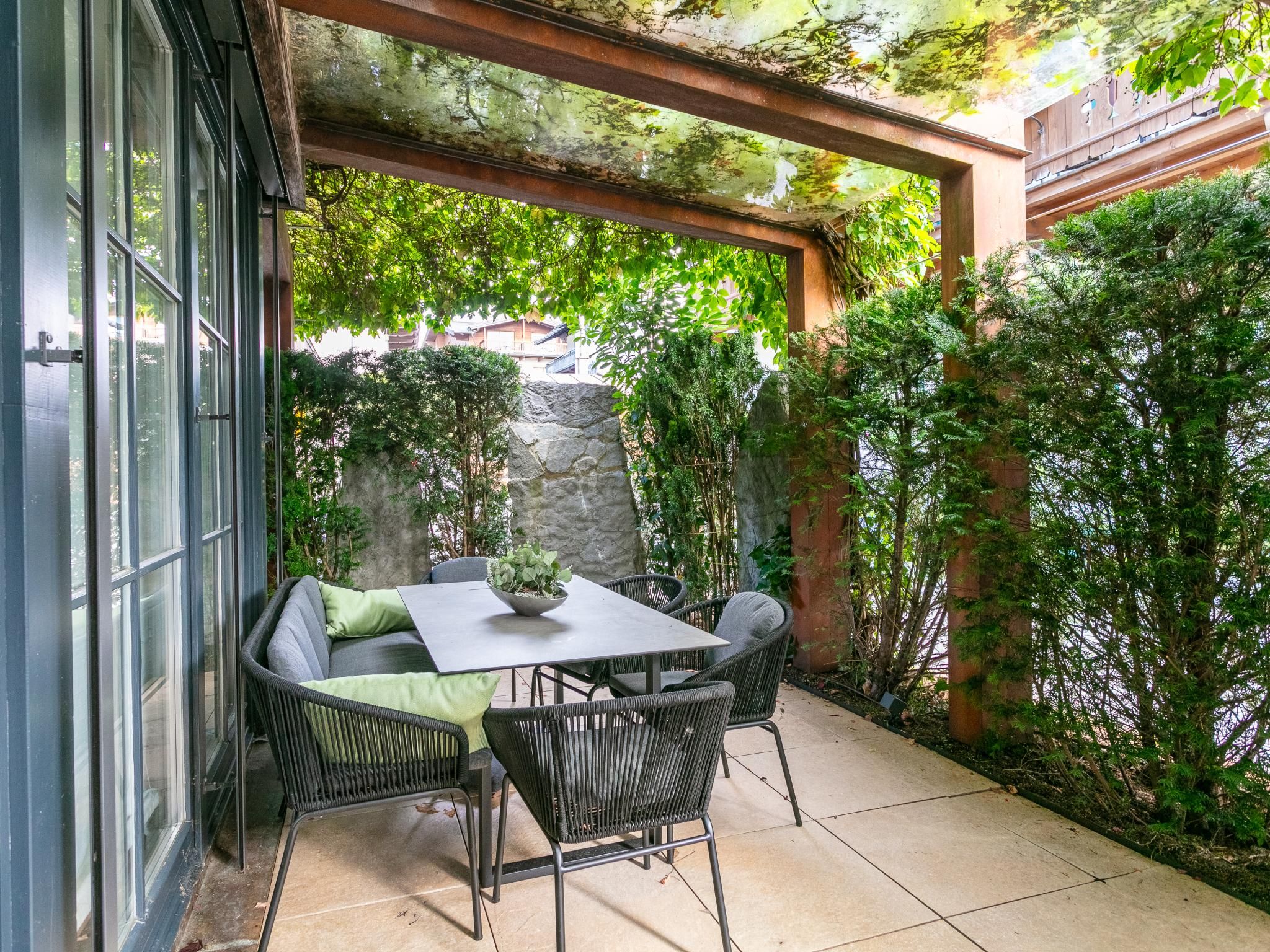 An outdoor patio with a table and chairs, surrounded by greenery and a stone wall.