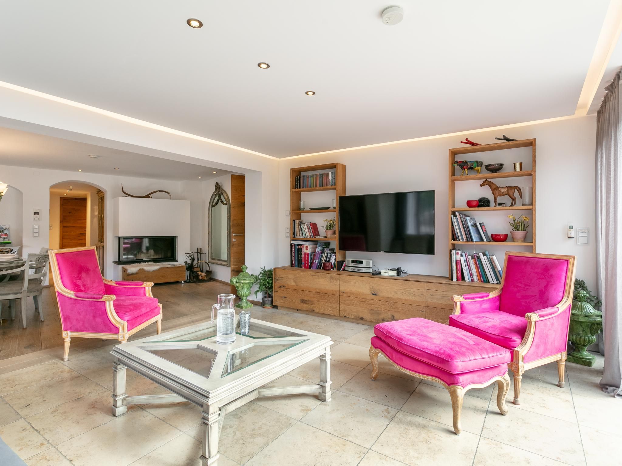 A modern living room with a flat-screen TV, two pink armchairs, a glass coffee table, and a wooden bookshelf.