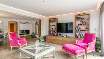 A modern living room with a flat-screen TV, two pink armchairs, a glass coffee table, and a wooden bookshelf.