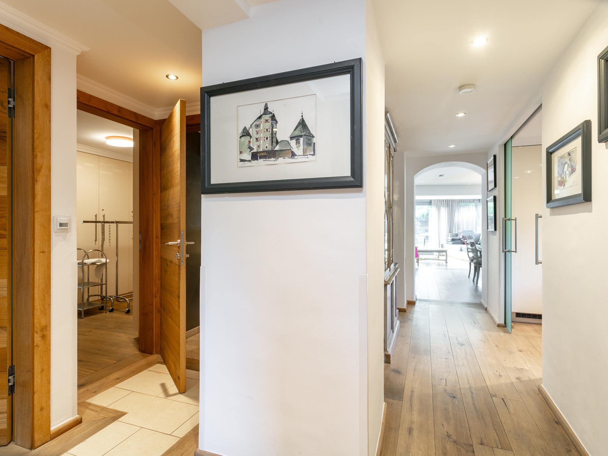 Um corredor com piso de madeira, um quadro pendurado na parede e uma porta que dá para um quarto com cama e mesa de cabeceira.