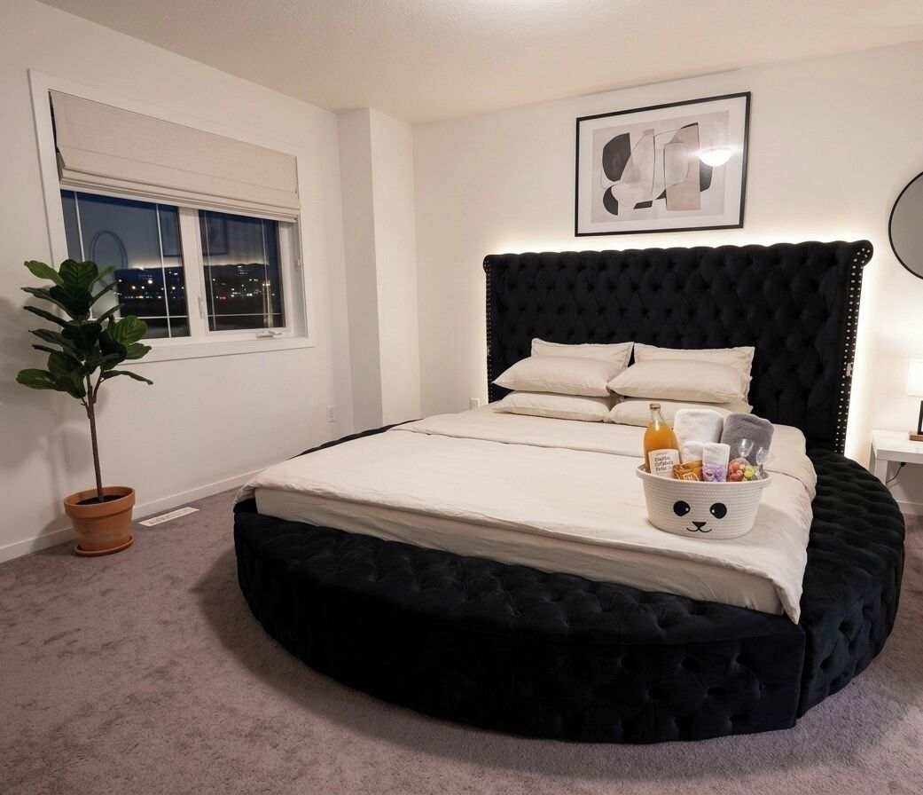 Quarto com cama redonda, janela, planta em vaso, quadro na parede e mesa de cabeceira com abajur.