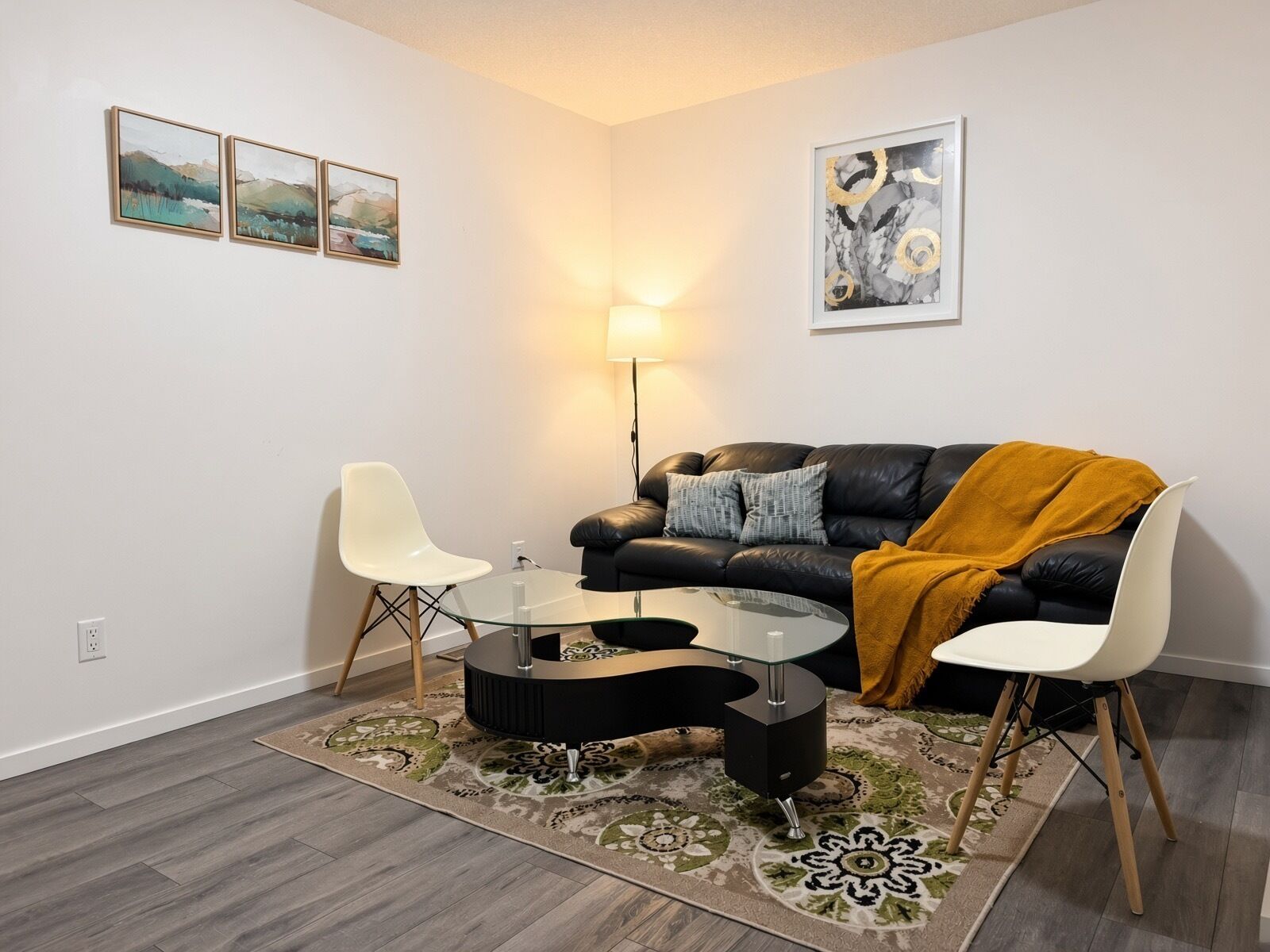 Een moderne woonkamer met een glazen salontafel, twee witte stoelen, een zwarte leren bank met een mosterdkleurig plaid, en een vloerkleed met een patroon.