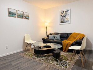 Un salon moderne avec une table basse en verre, deux chaises blanches, un canapé en cuir noir agrémenté d’un plaid jaune moutarde et un tapis à motifs.
