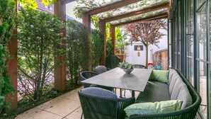 Eine Terrassenanlage im Freien mit Tisch und Stühlen, umgeben von Grünpflanzen und einem Pergolen-Aufbau.