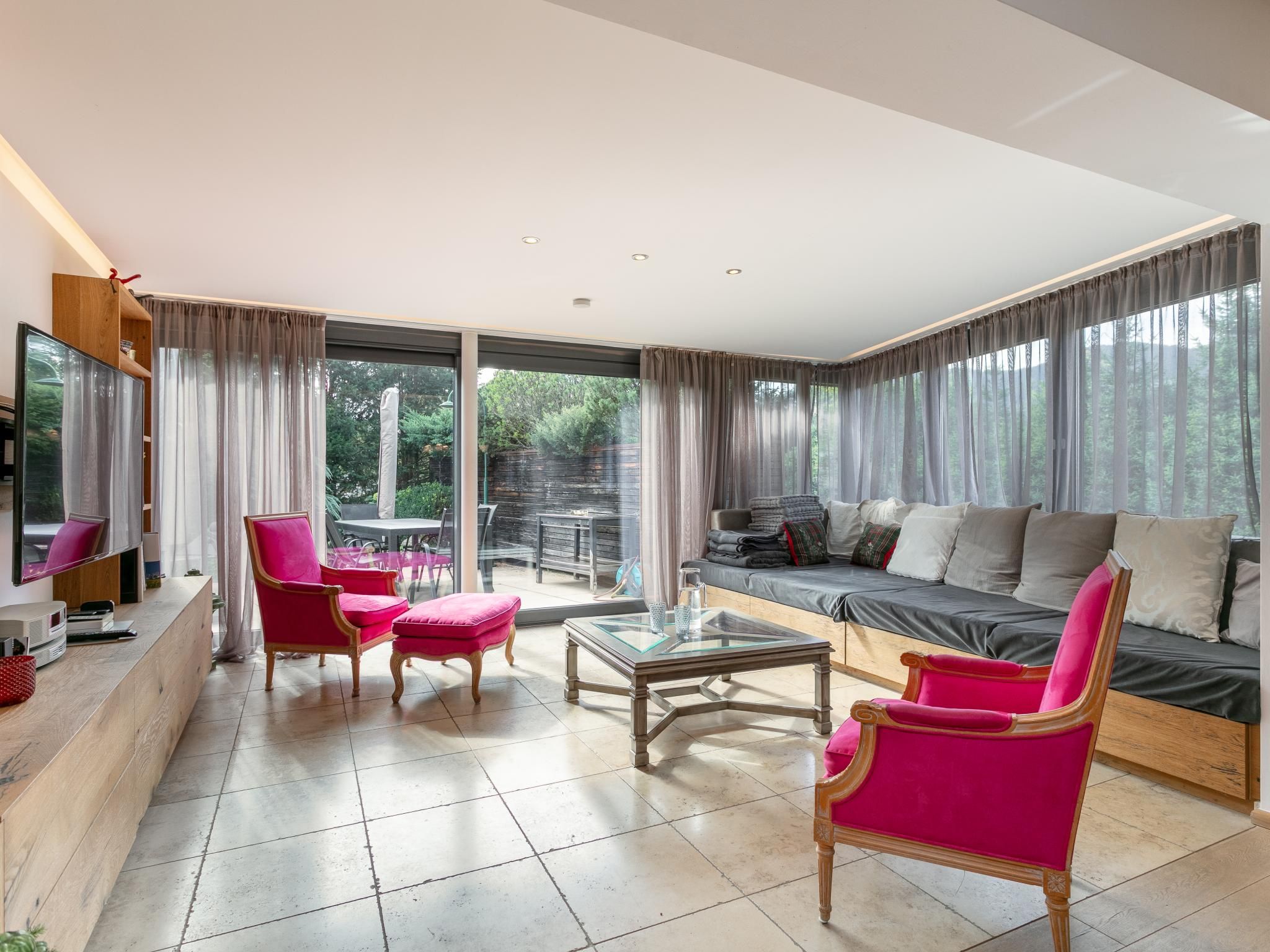 Een ruime woonkamer met een grote bank, een salontafel en twee fauteuils. Er staat een televisie op een houten dressoir en de kamer heeft grote ramen met gordijnen.