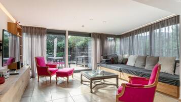 Een ruime woonkamer met een grote bank, een salontafel en twee fauteuils. Er staat een televisie op een houten dressoir en de kamer heeft grote ramen met gordijnen.