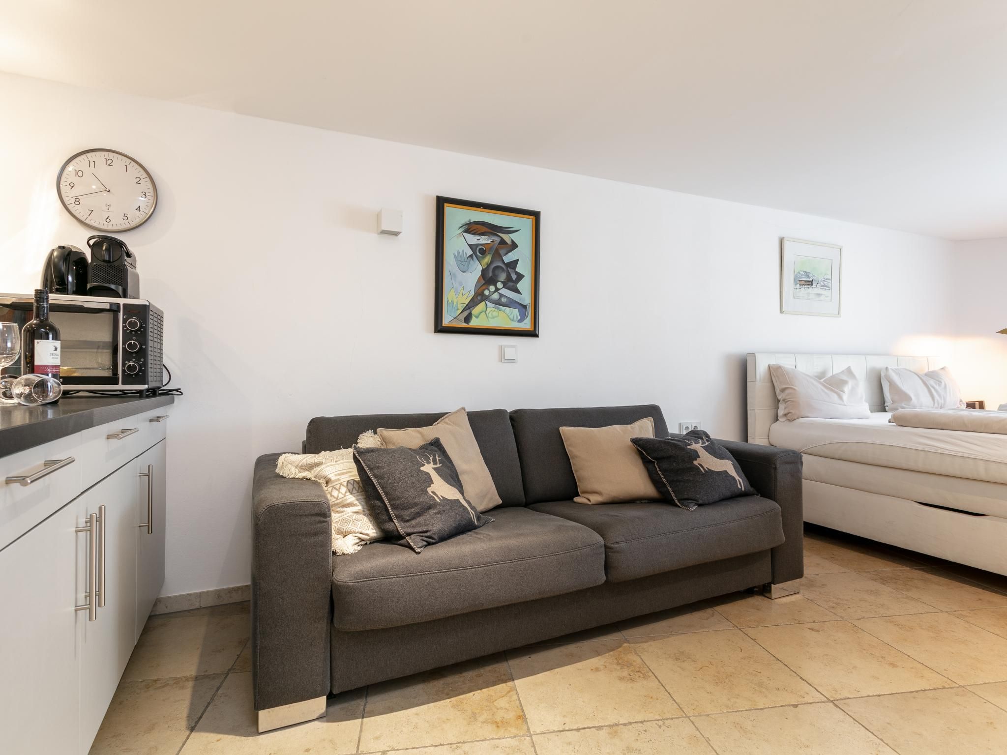 Een moderne woonkamer met een grijze bank, een wit bed, een keuken met een magnetron, en een wandklok.
