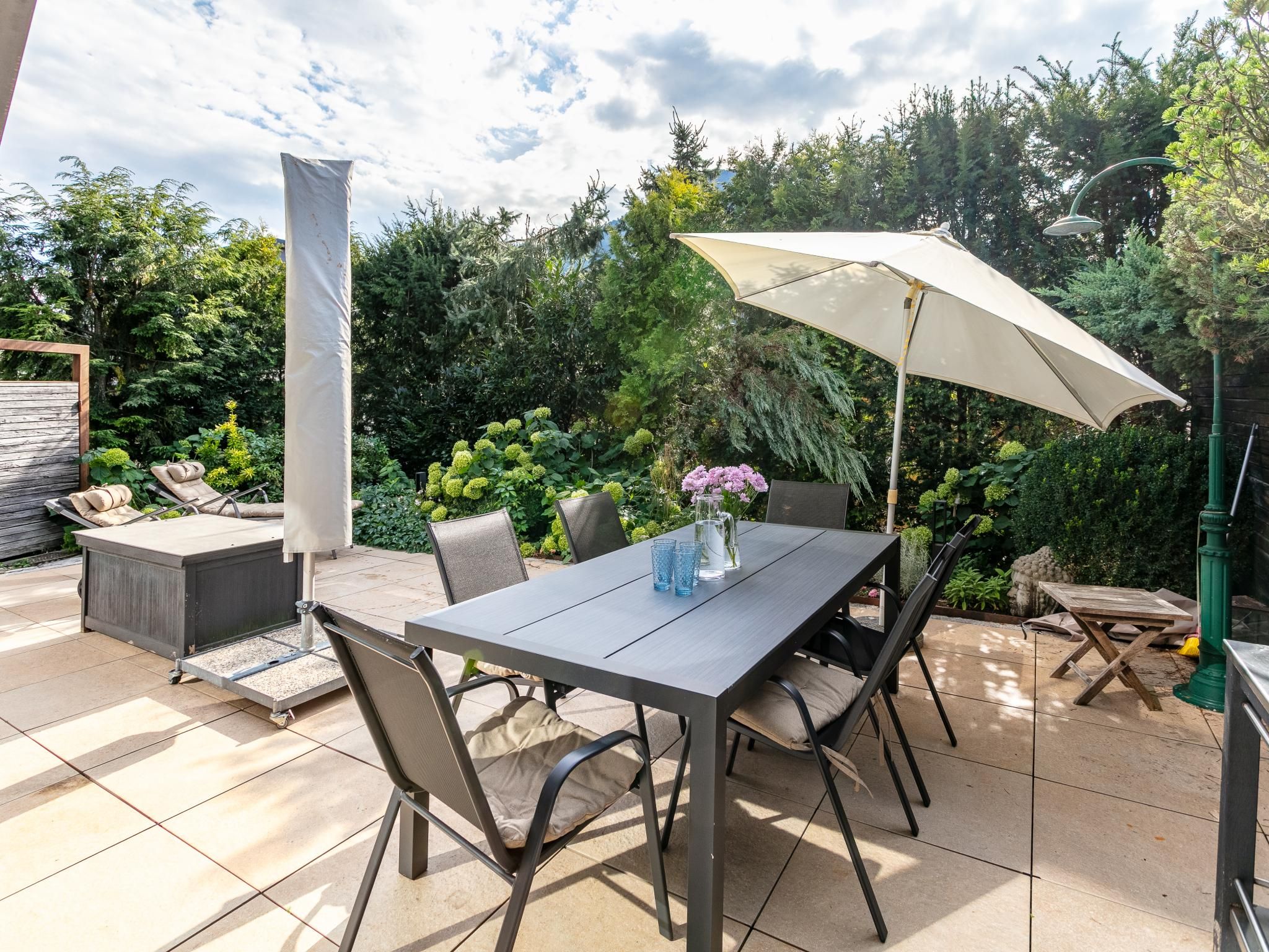 Un patio al aire libre con una mesa, sillas, una sombrilla y un jardín al fondo.