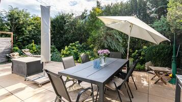Un patio al aire libre con una mesa, sillas, una sombrilla y un jardín al fondo.