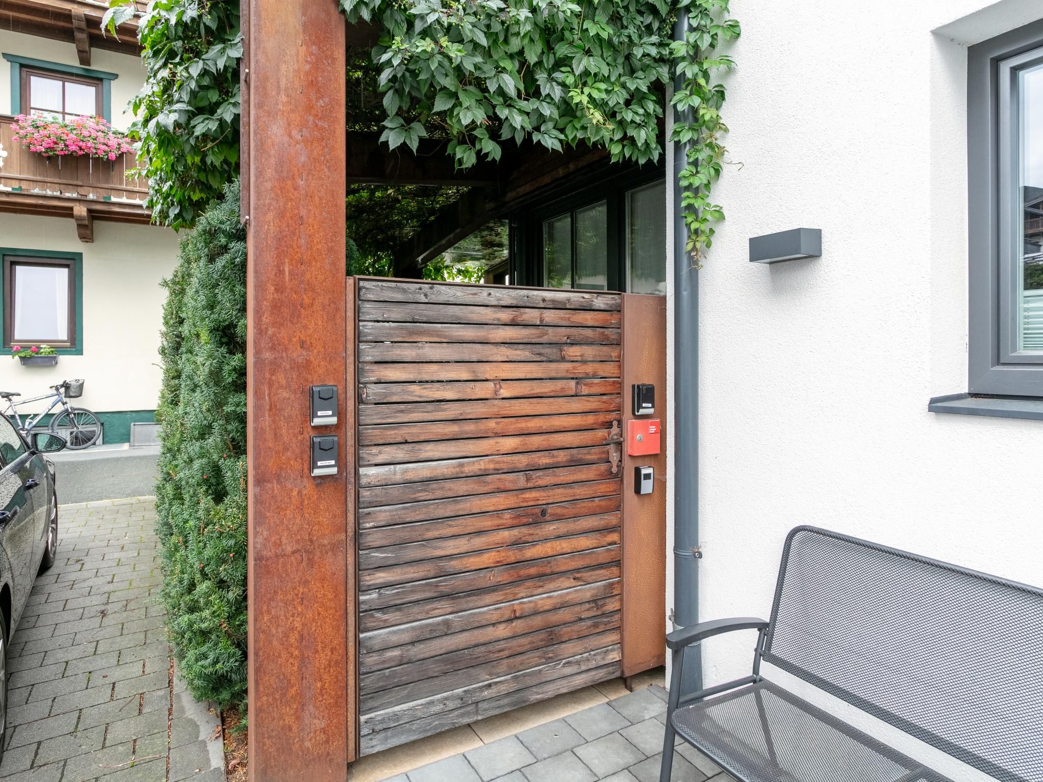 Een houten poort met een metalen handgreep en een rode noodknop, een bankje en een fiets die er staan.